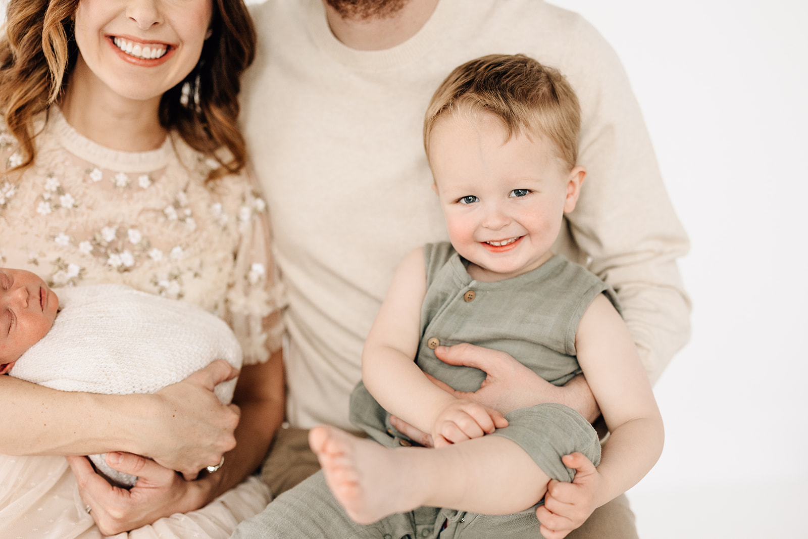 A young toddler smiles in a green onesie while sitting in dad's lap next to mom holding a sleeping newborn in a studio after visiting Baby Boutiques St. Louis