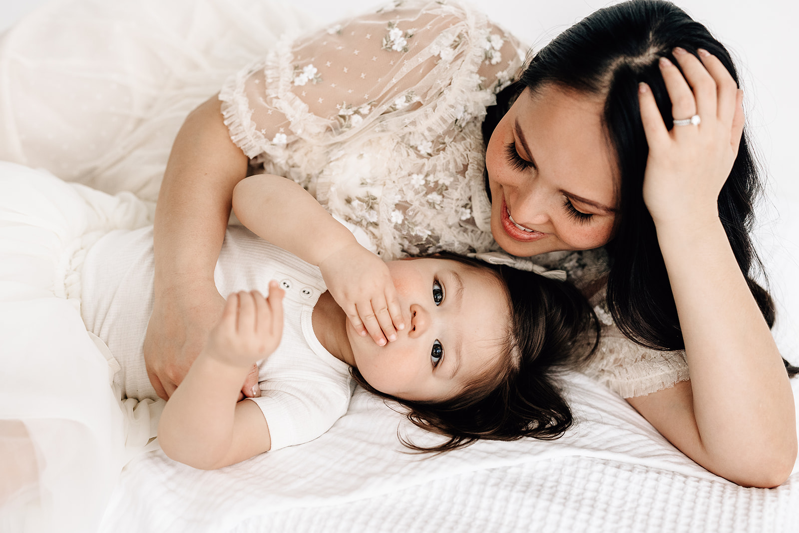 A mother in a white lace embroidered dress lays on a bed cuddling with her toddler daughter thanks to Fertility Acupuncture St Louis