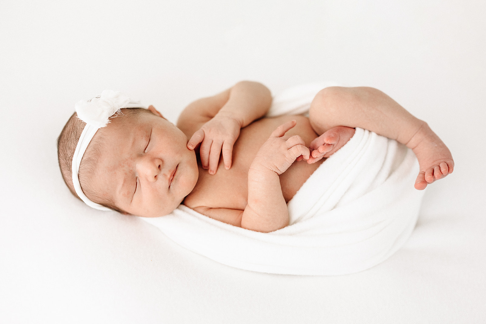 A newborn baby girl sleeps in a white half swaddle and bow