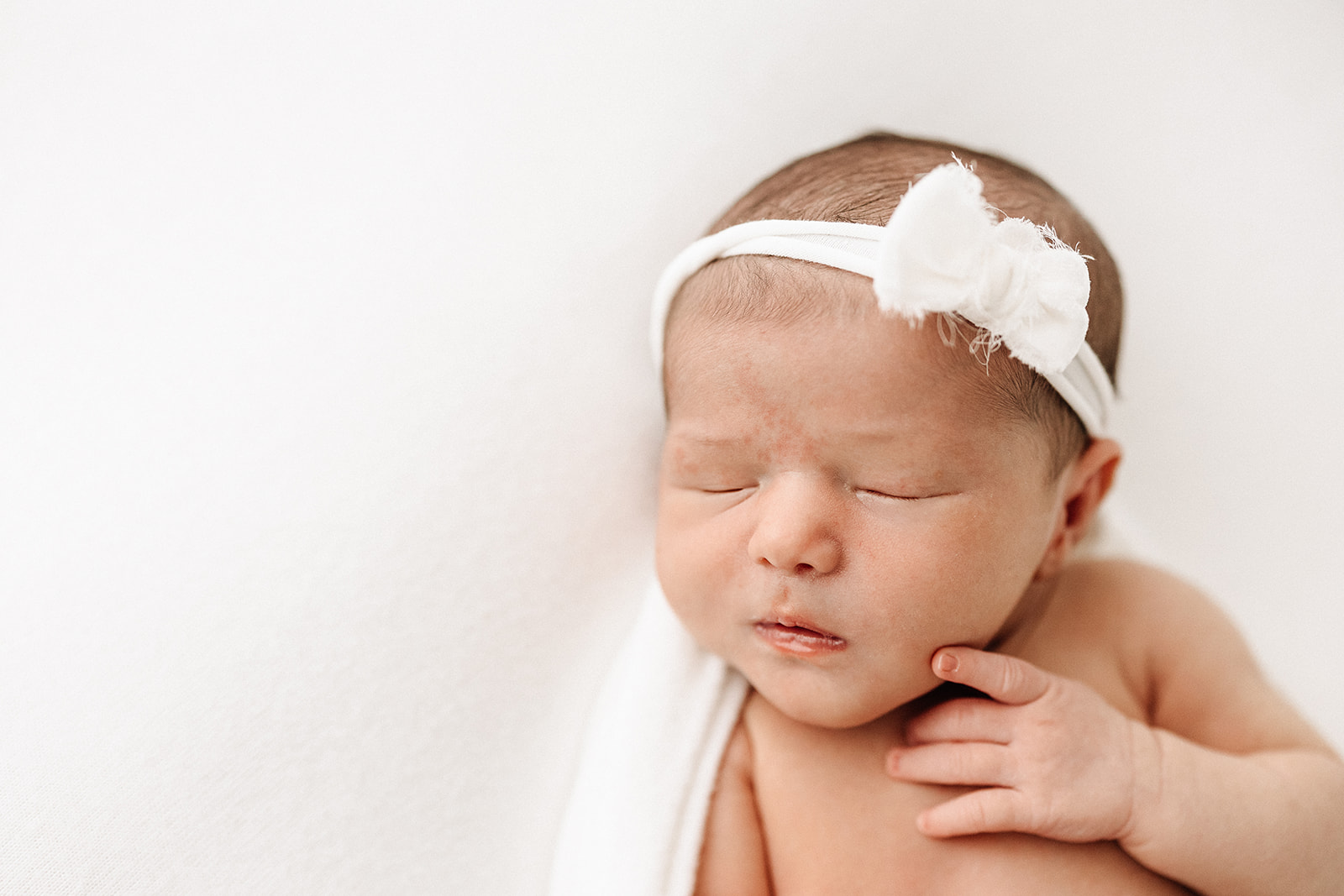 A newborn baby sleeps in a white bow headband in a studio before some ISR St. Louis