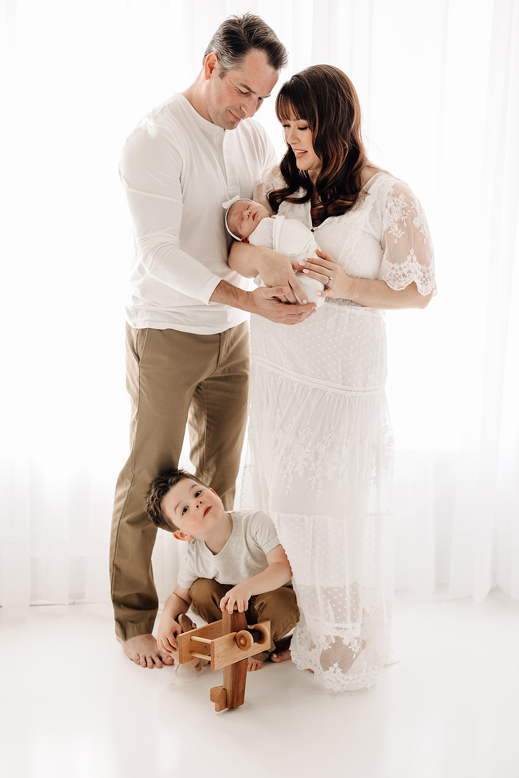 Happy mom and dad stand in a studio smiling at their newborn baby in mom's arms while their son plays below them before visiting playgrounds St. Louis