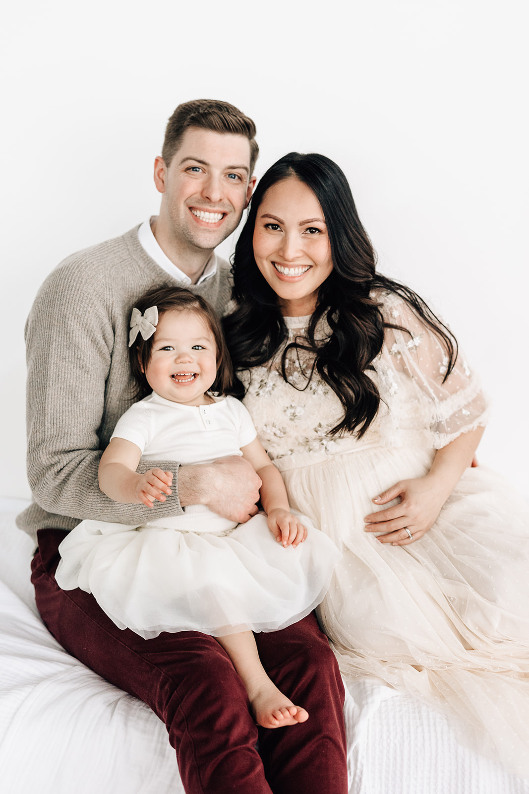 A happy pregnant mother sits on a bed in a white dress with her husband and their toddler daughter in his lap