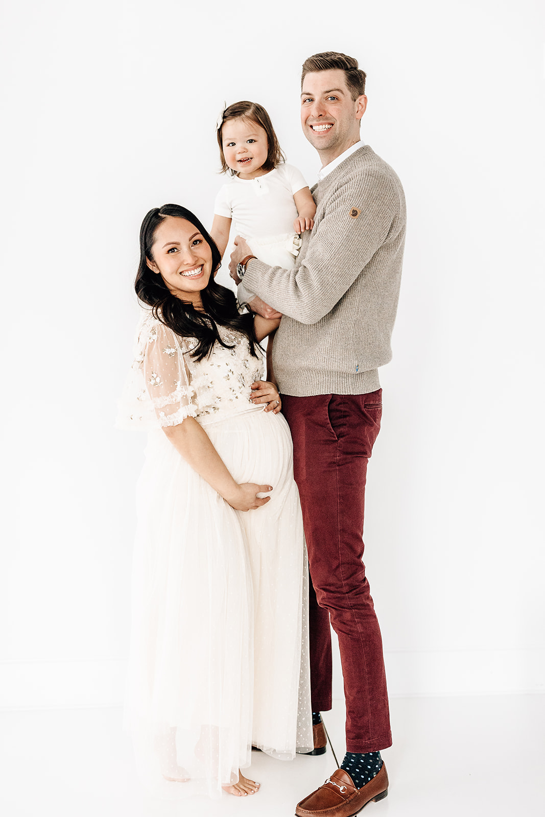 Happy mom and dad stand in a studio while dad holds their smiling toddler and mom holds her pregnant bump after visiting Preschools in St. Louis