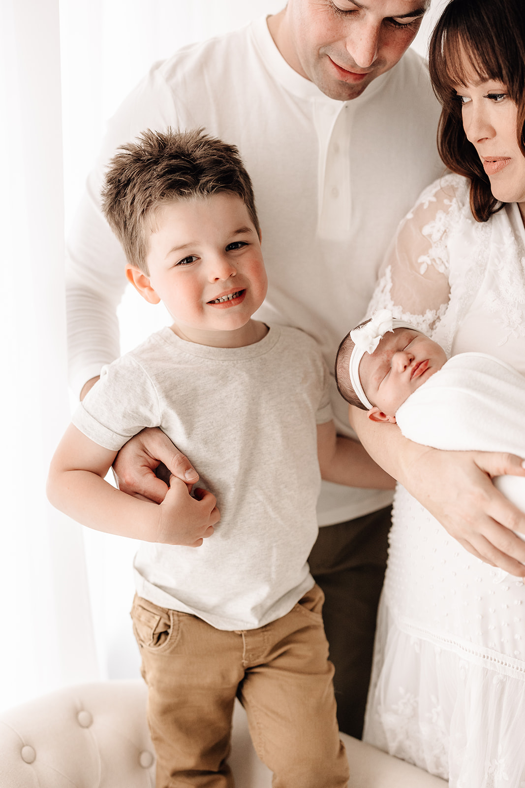 A happy toddler boy stands with mom and dad as they hold his sleeping newborn baby sister