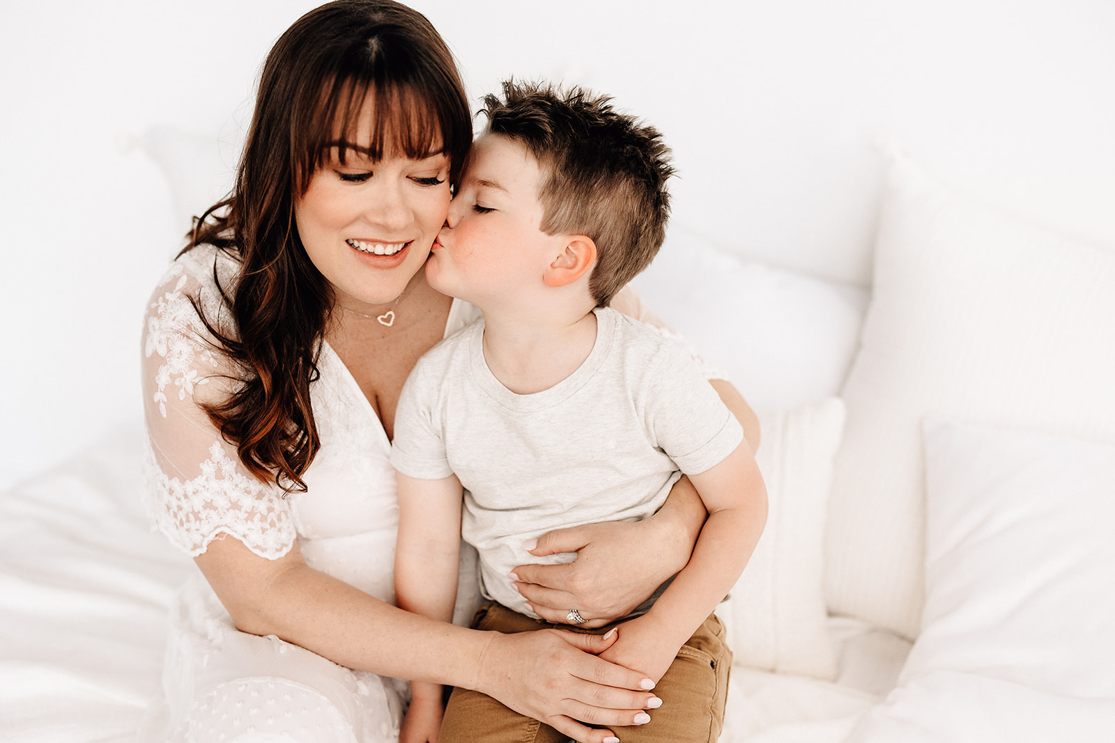 A mother in a white dress is kissed by hers toddler son as he sits in her lap on a bed before exploring splash pads St. Louis