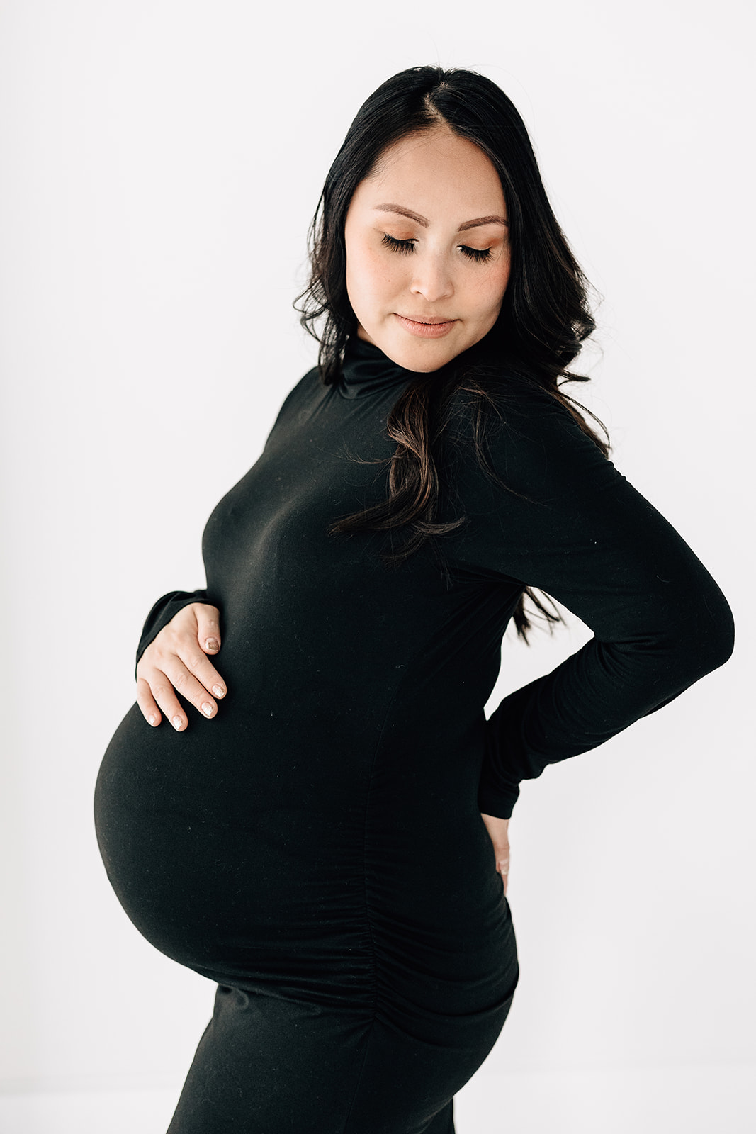A mother to be smiles down her shoulder while standing in a studio in a black maternity gown