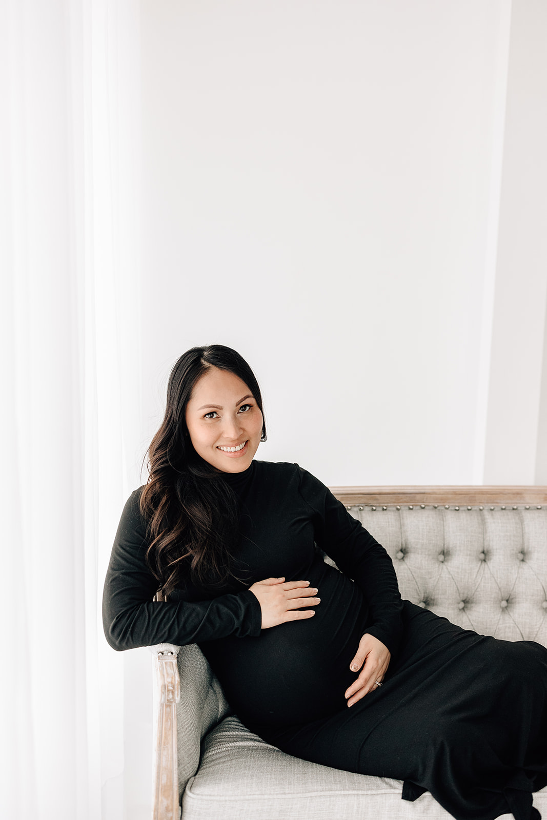 An expecting mother wearing a long black maternity gown smiles while sitting across a bench in a studio after meeting Anna The Doula