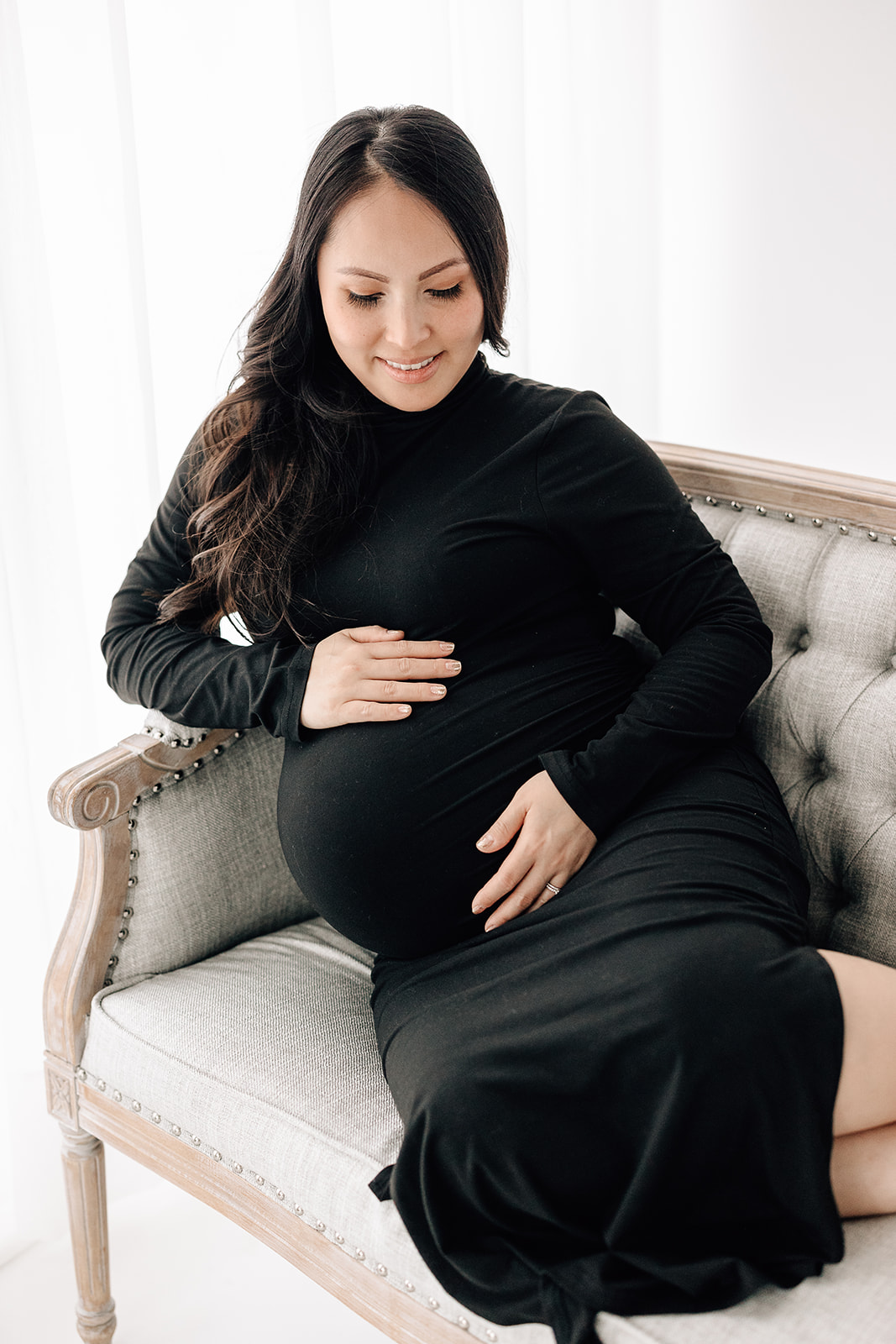 A mom to be in a black maternity gown sits across a couch while smiling down at her bump after meeting Anna The Doula