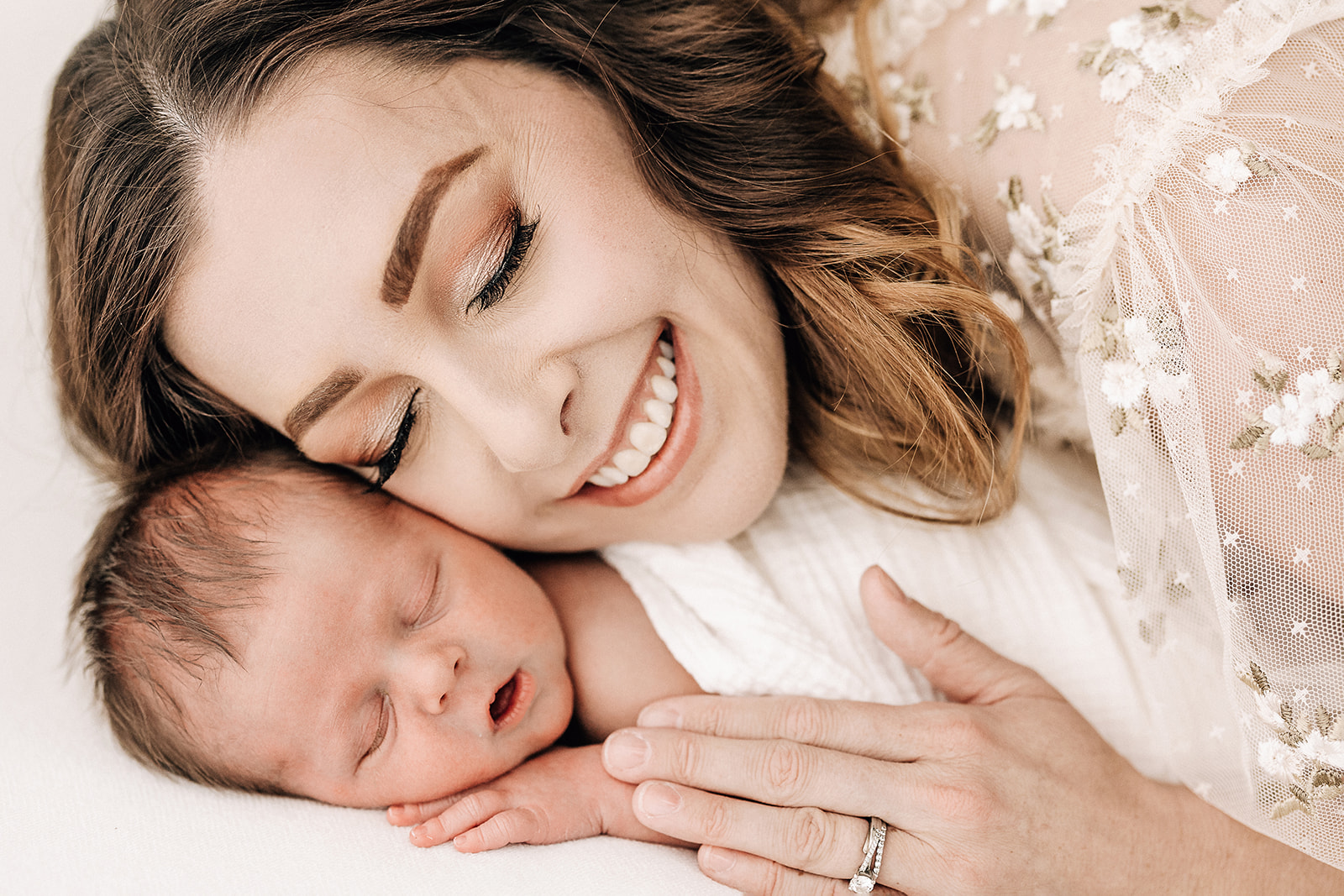 A smiling mom snuggles with her sleeping newborn baby on a white bed after visiting Be By Baby