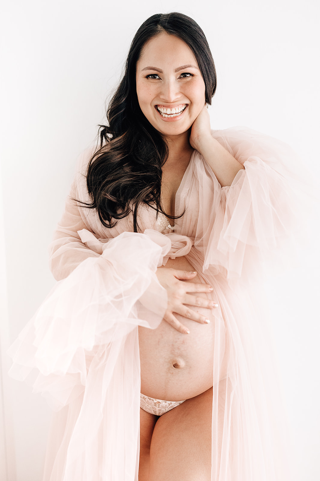 A laughing pregnant woman stands in a studio with a hand on her bump in an open pink tule maternity gown