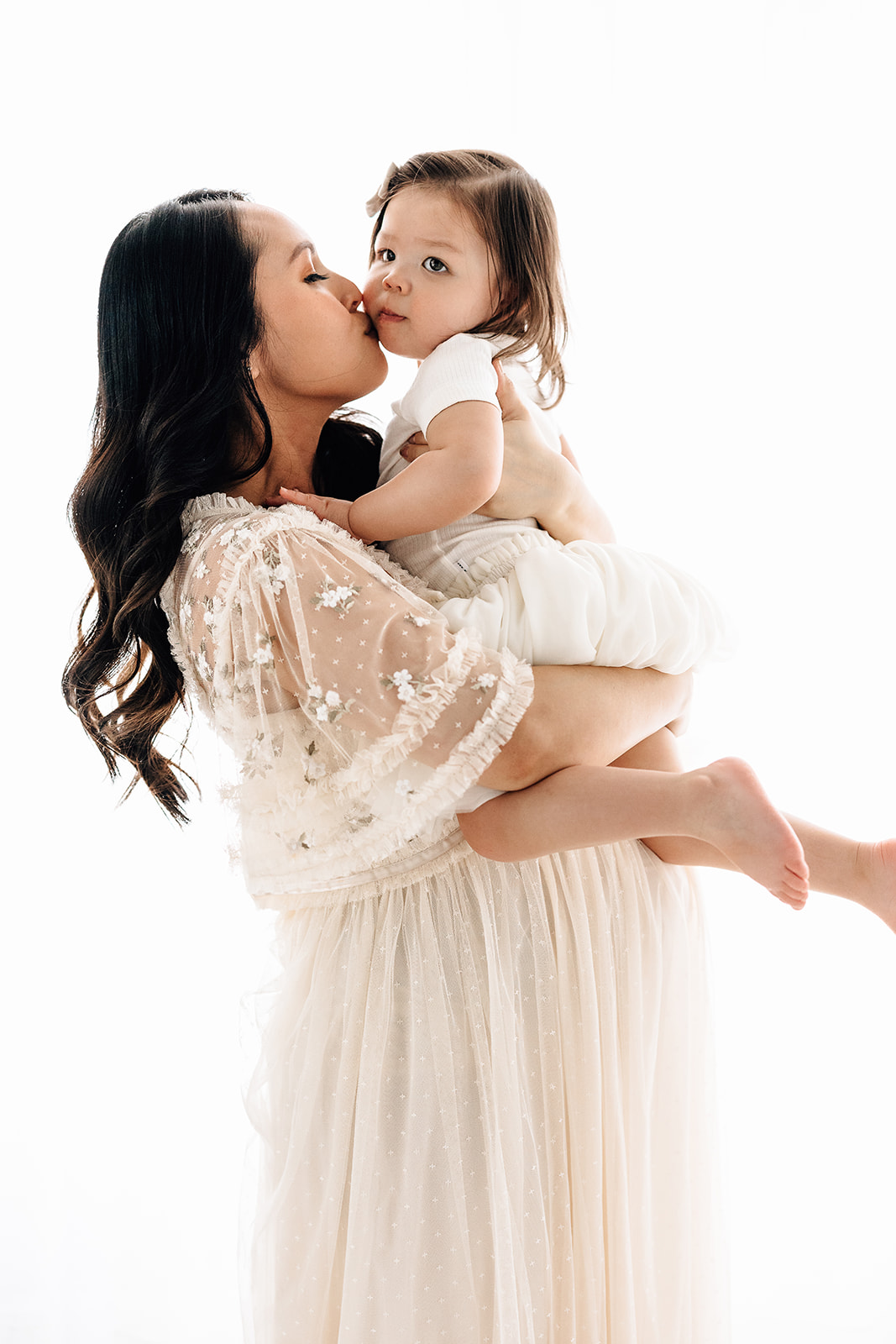 A mother kisses her toddler daughter in a white dress while holding her in a studio
