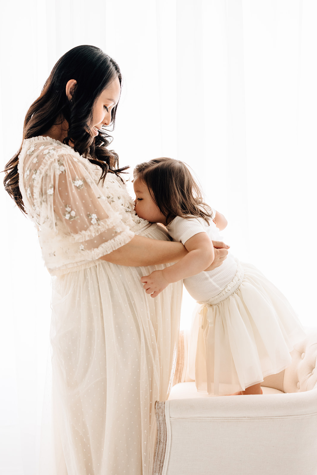 A toddler girl stands on a chair to kiss her mom's pregnant bump in a studio after visiting TLC Family Care