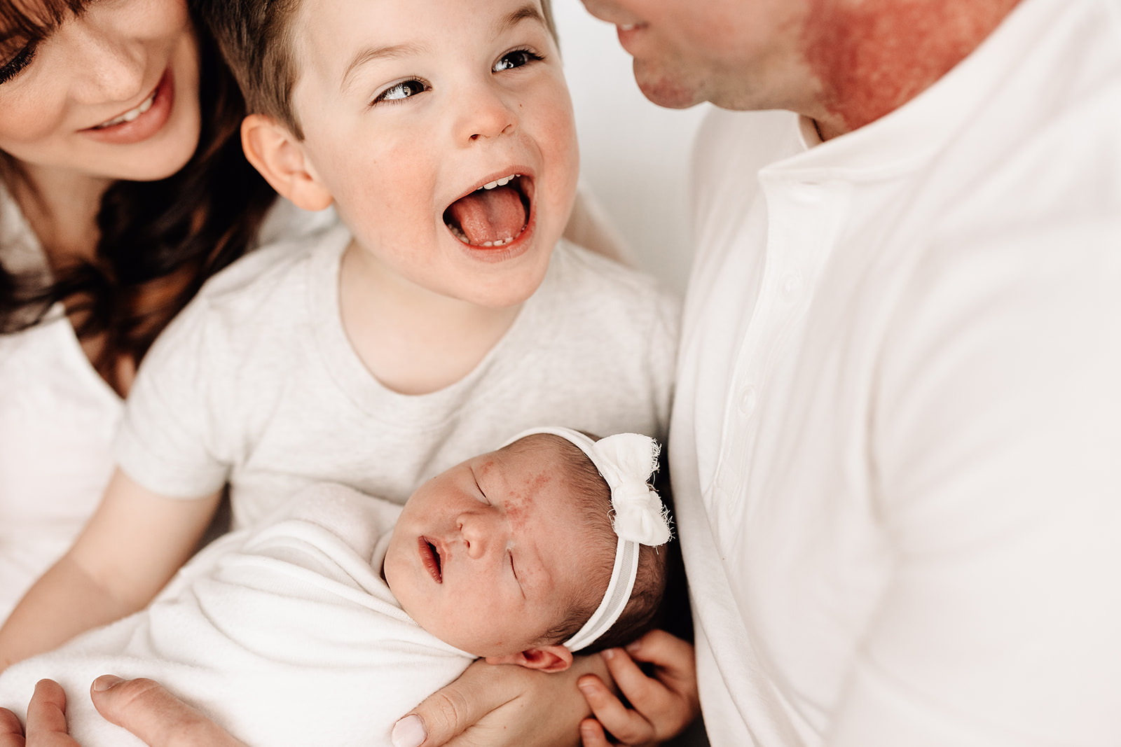 A toddler boy laughs while holding his sleeping newborn baby sister in dad's lap