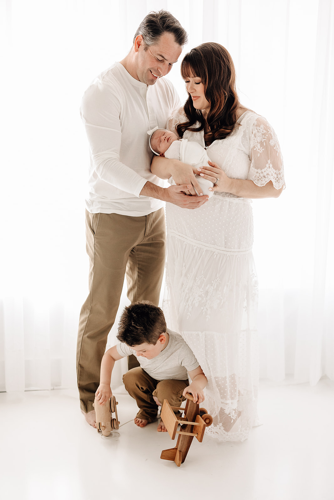 Happy mom and dad cradle their sleeping newborn daughter while standing above their son playing with wooden toys after meeting Gateway Nanny Agency