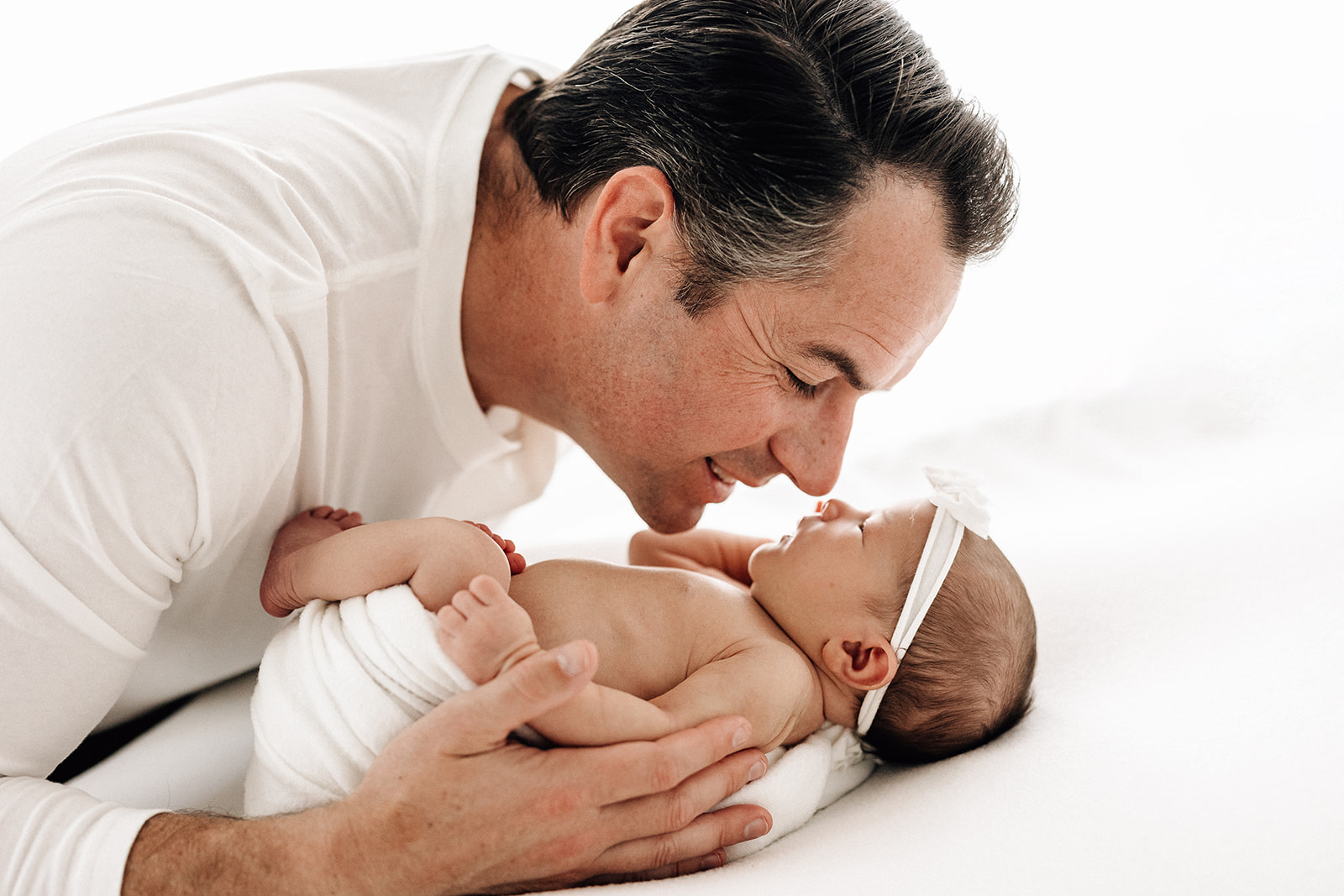 A happy father snuggles his newborn daughter in a white swaddle on a bed before meeting Gateway Nanny Agency