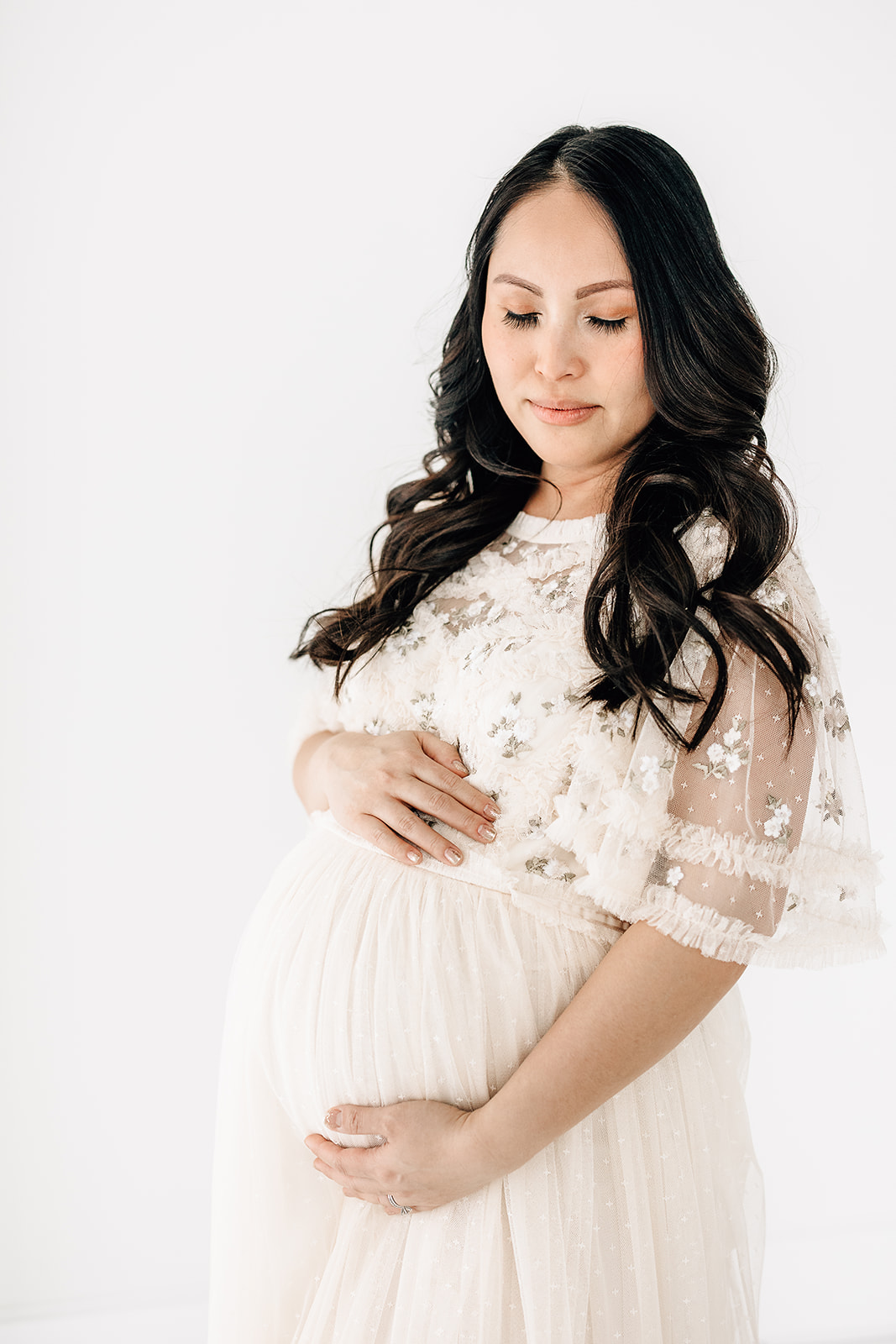 A pregnant woman in a white lace maternity gown holds her bump while standing in a studio after visiting Missouri Baptist Birthing Center