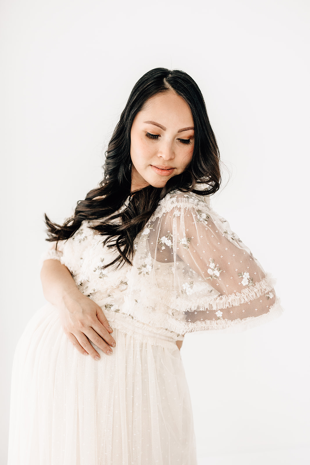 A mother to be in a lace maternity gown stands in a studio with a hand on her belly and back after visiting Missouri Baptist Birthing Center