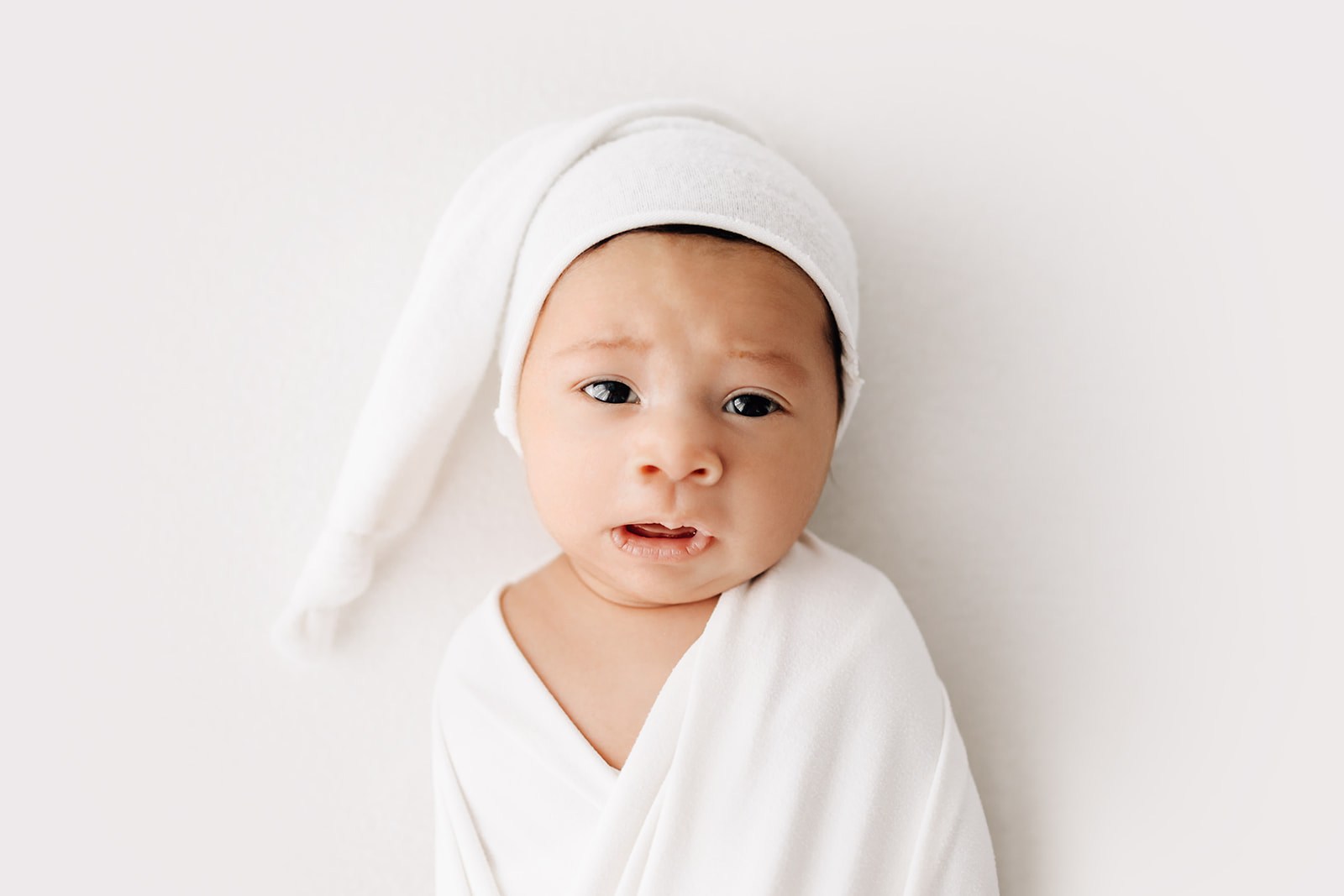 A newborn baby lays on a white bed in a white blanket and sleep cap after meeting Tiny Transitions Sleep Consulting