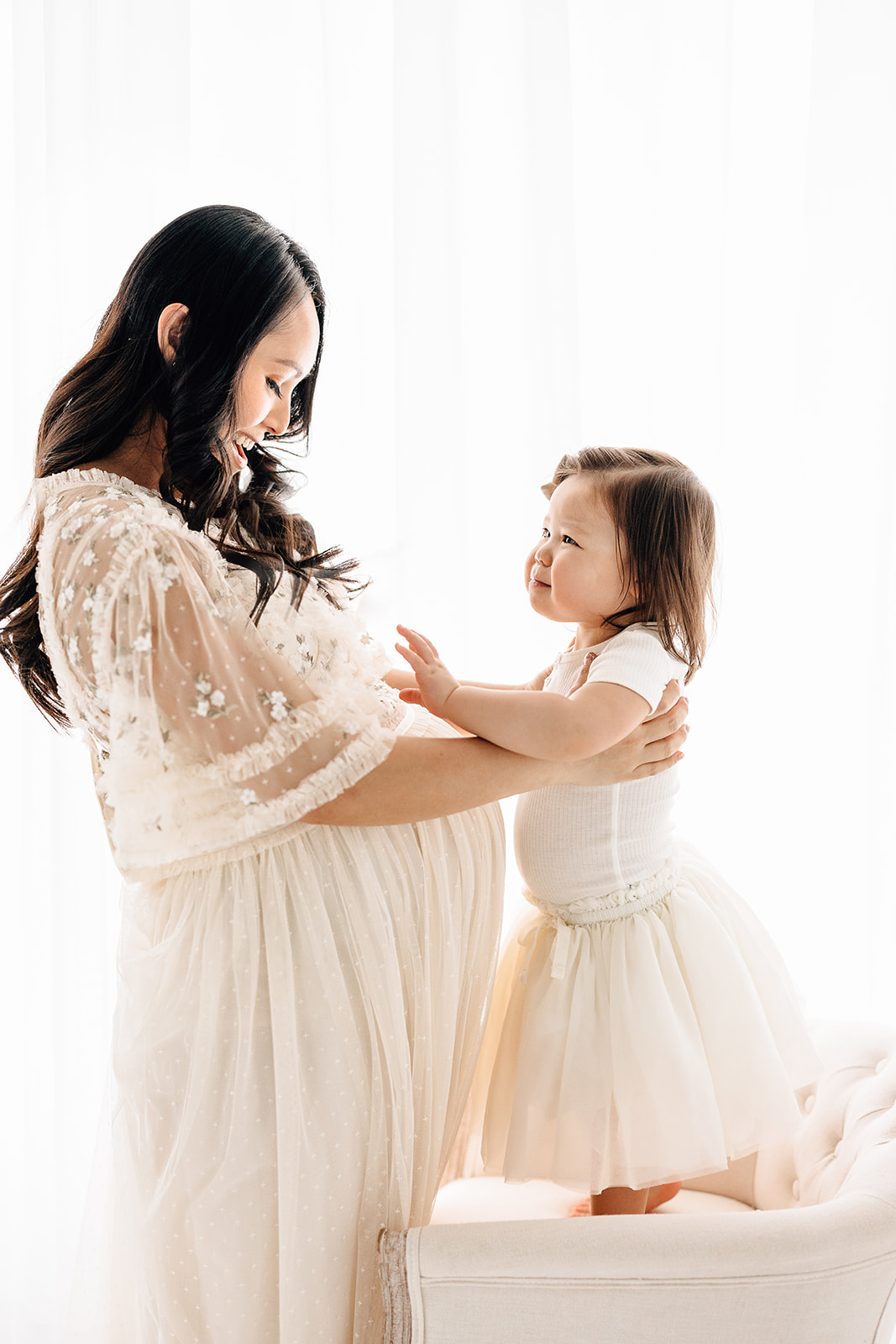 A toddler girl reaches for mom while standing on a chair in a studio window