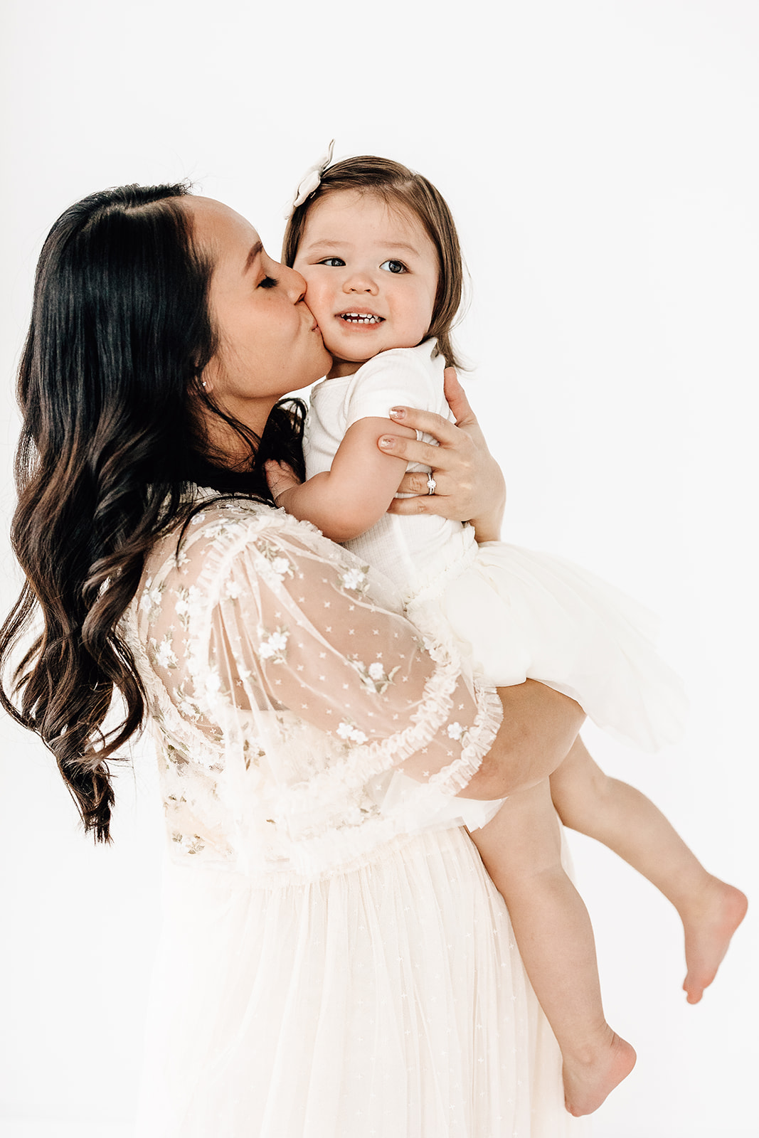 A happy toddler girl sits in mom's arms while being kissed on the cheek in a white dress before visiting Imagination toys St. Louis