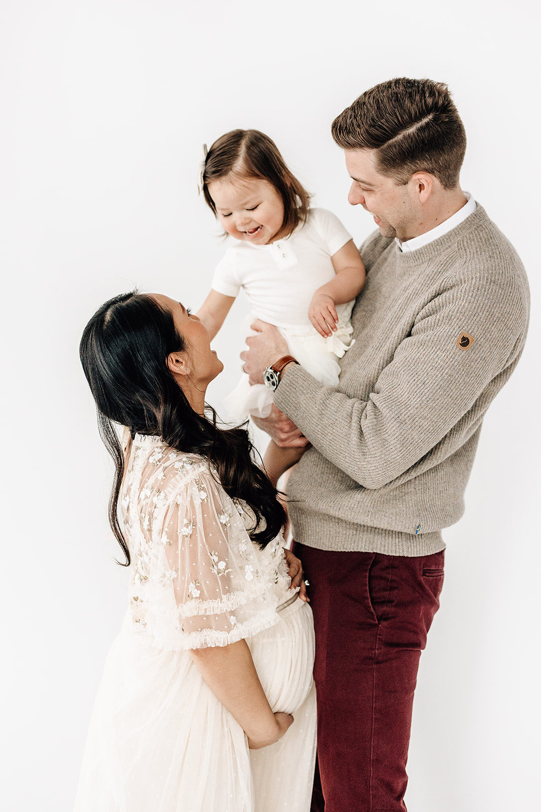 A happy toddler girl in a white dress plays with pregnant mom while sitting in dad's arms before visiting Imagination toys St. Louis