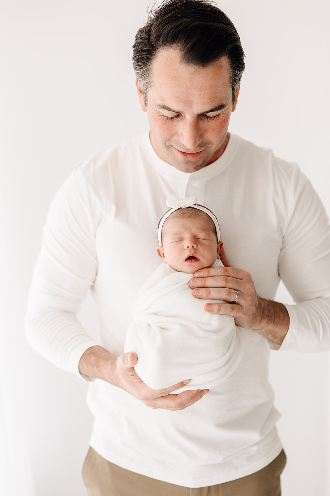 A newborn baby sleeps in dad's hands against his chest as he stands in a studio