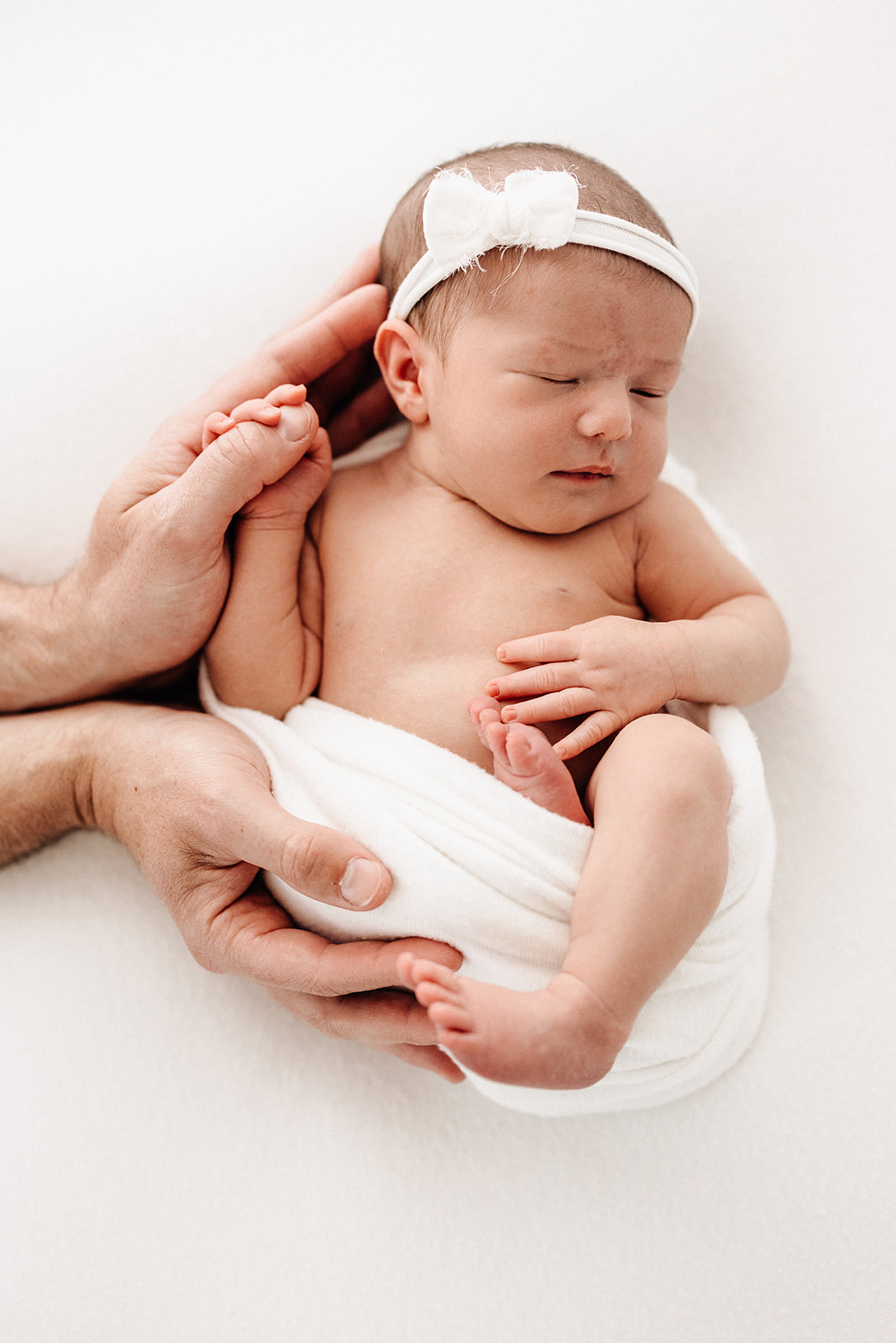 A newborn baby sleeps while holding dad's thumb on a white bed with a matching headband