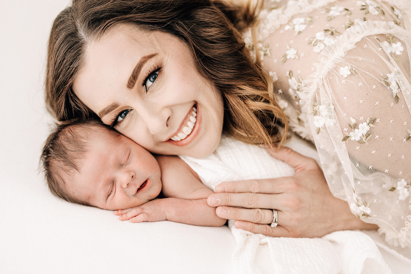 A happy new mom snuggles with her sleeping enwborn baby on a white bed