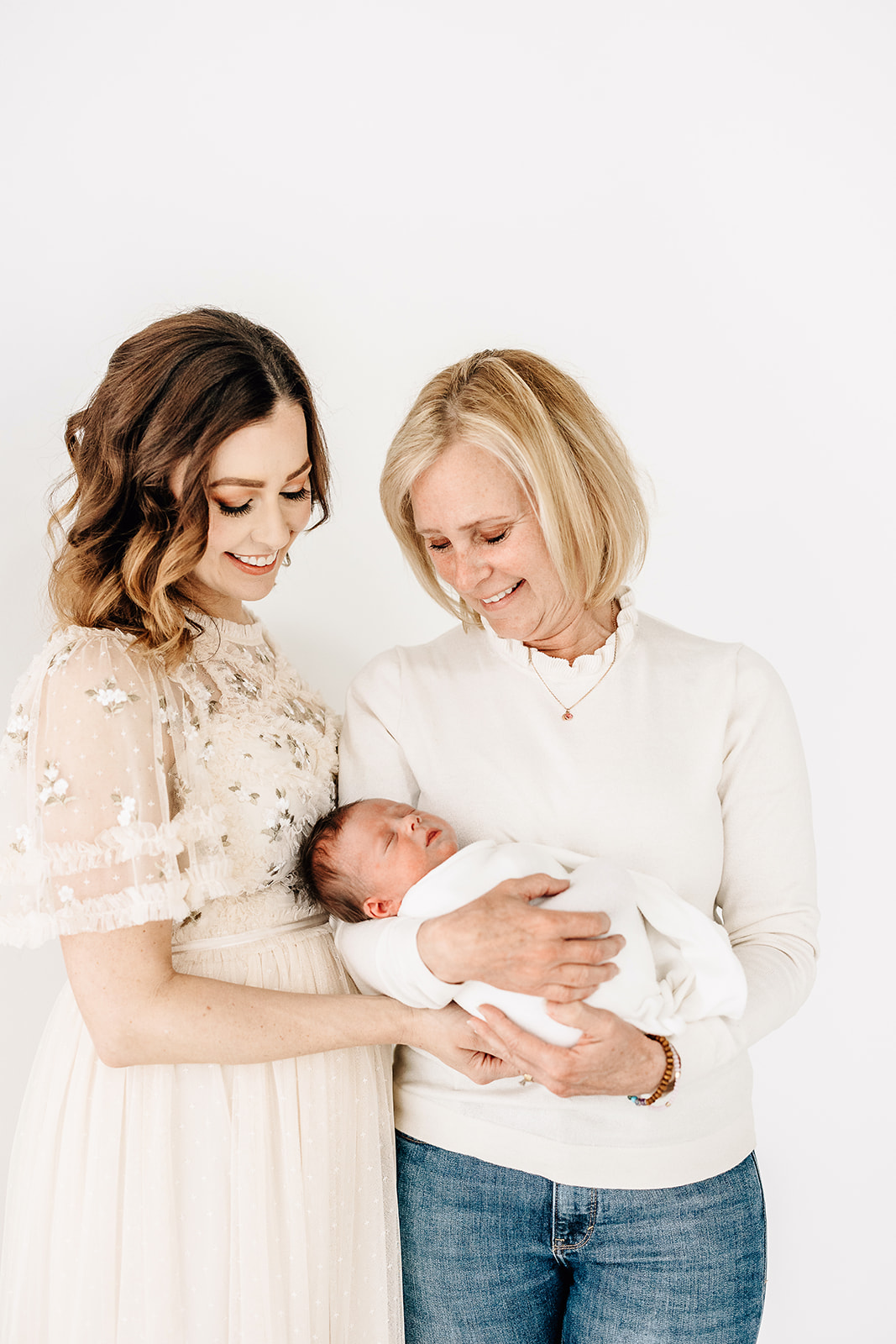 A new grandmother stands holding her sleeping newborn baby with mom in a studio after meeting Sleepyhead Solutions