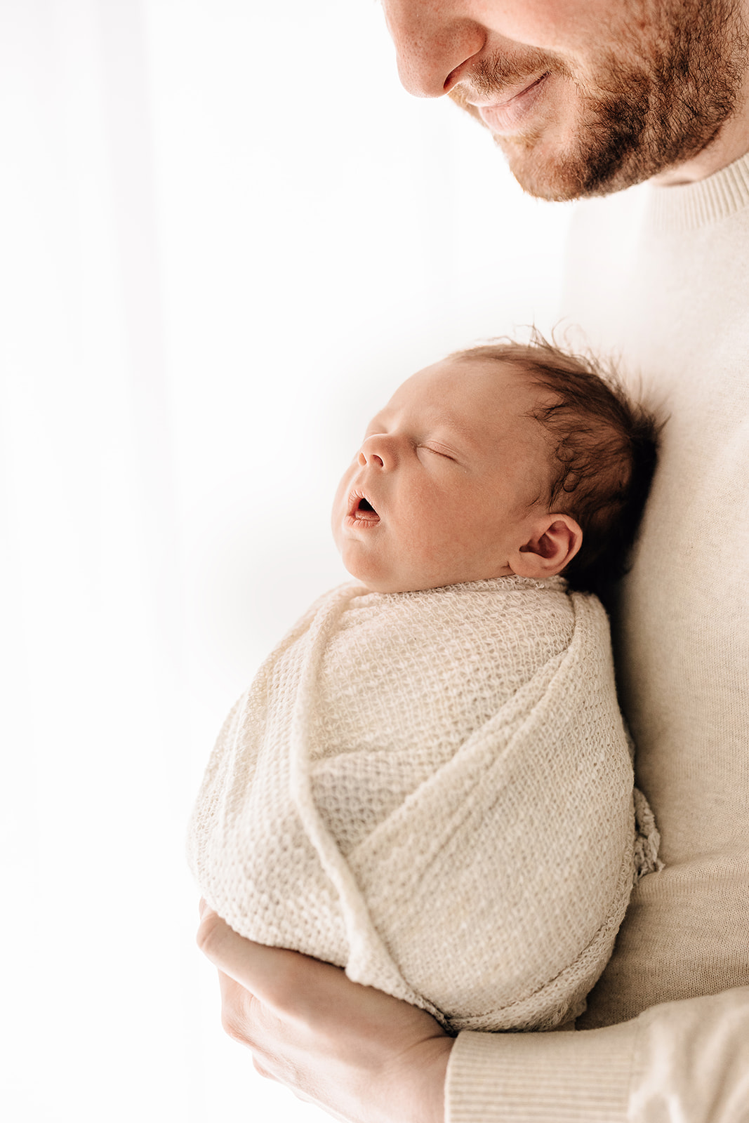 A newborn baby sleeps against dad's chest in a window thanks to Sleepyhead Solutions