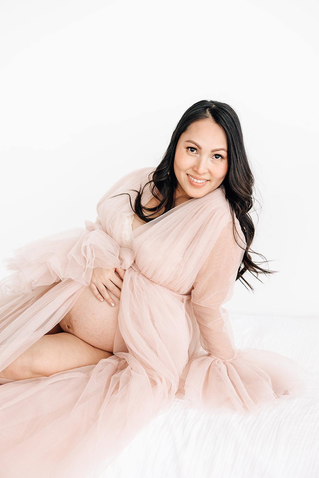 A mom to be sits across a white bed in a studio while resting a hand on her bump