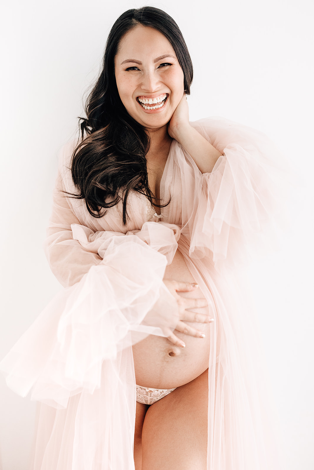 A laughing mom holds her bump while standing in a studio in a pink tule maternity gown after visiting St. Lukes Hospital Birthing Center