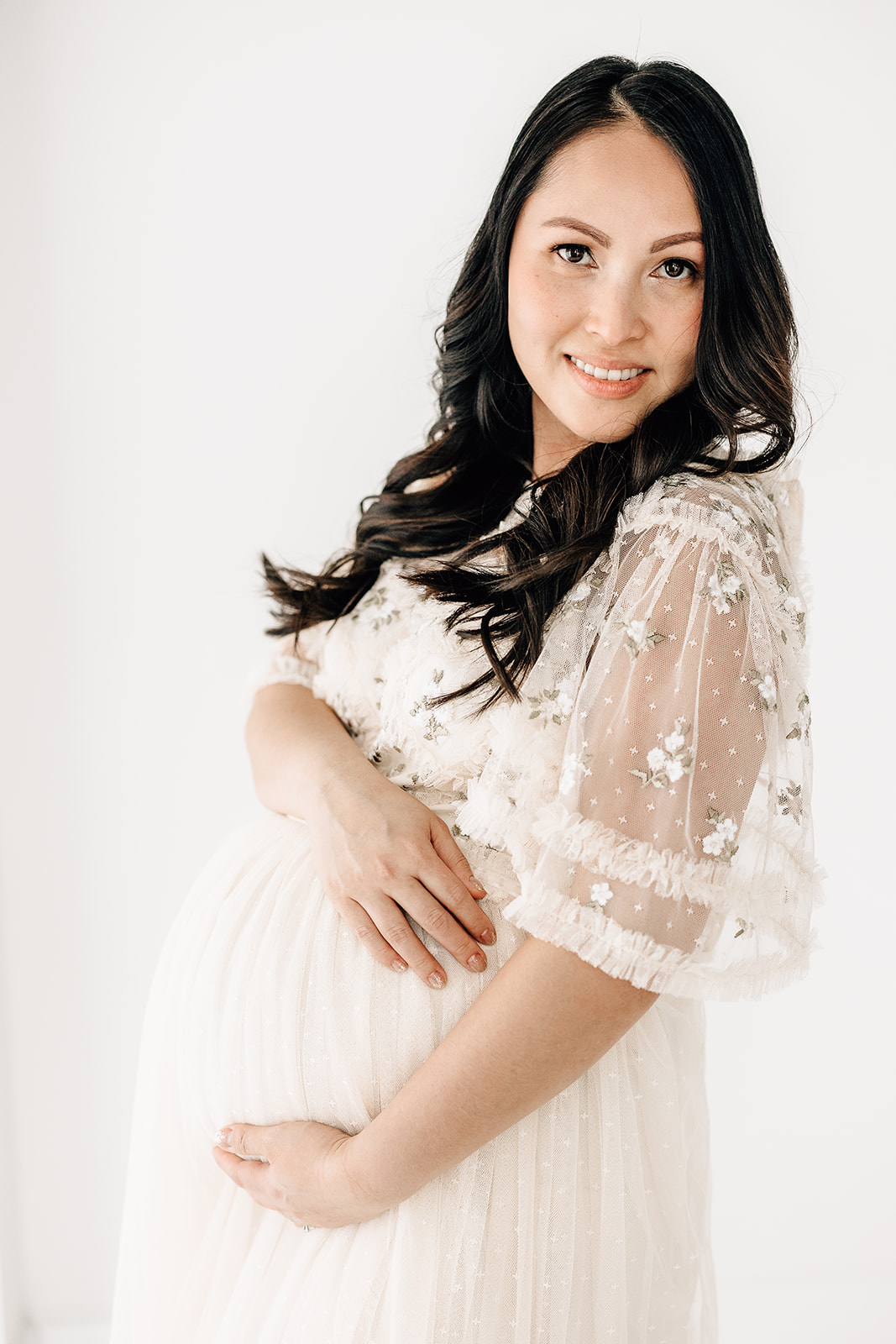 A happy mom to be in a white lace dress holds her bump while leaning on a wall in a studio after visiting St. Lukes Hospital Birthing Center