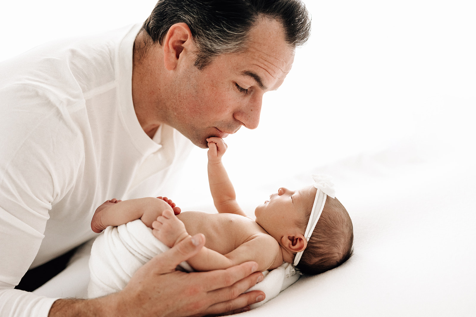 A newborn baby lays on a bed reaching up to dad's chin