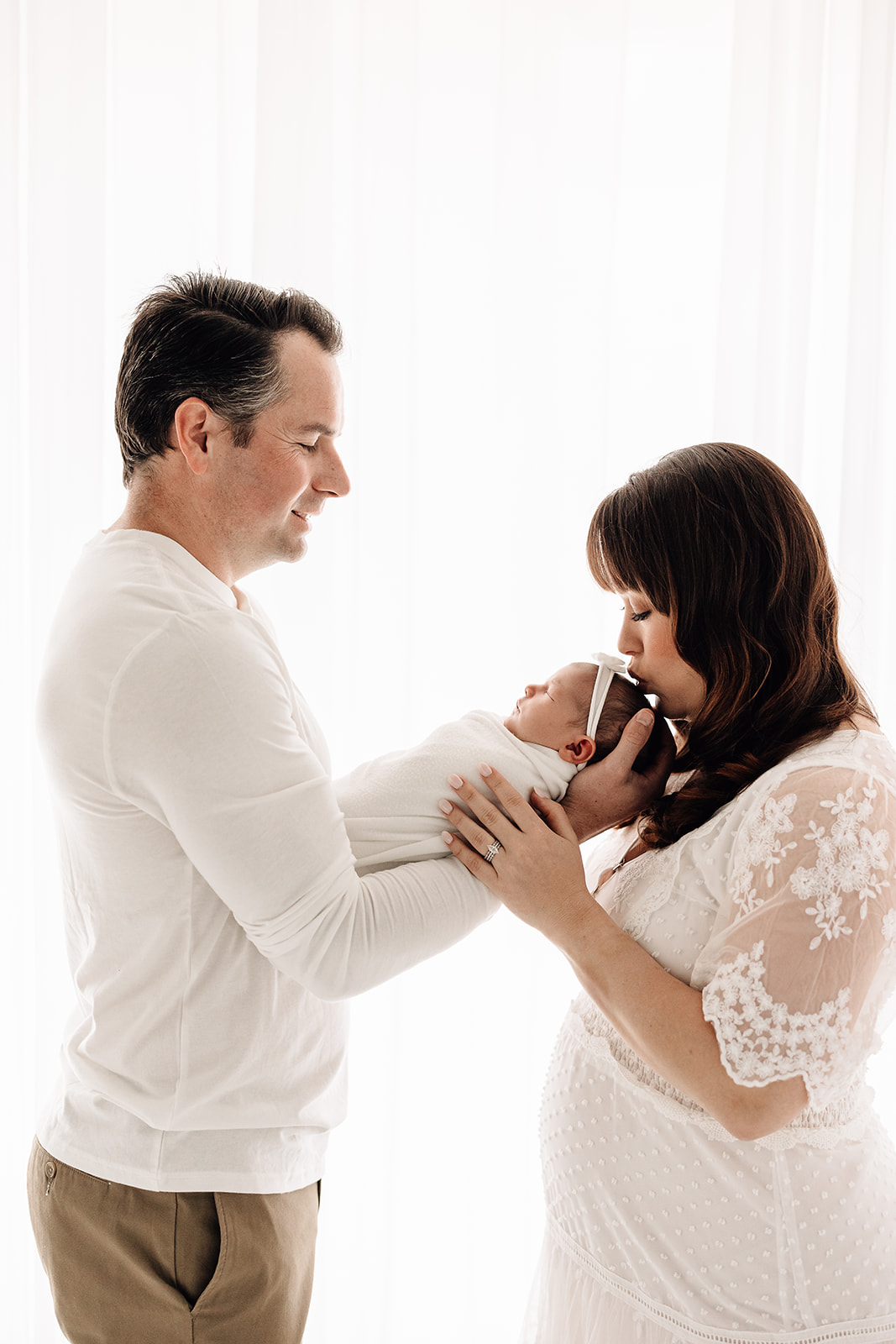 A new father holds his sleeping newborn baby out for mom to kiss while standing in a window before visiting The Magic House St. Louis