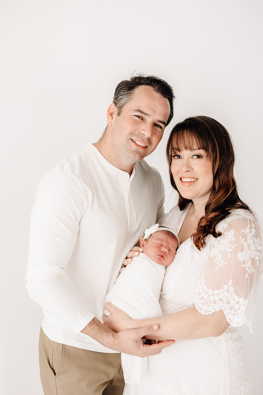 Happy new parents stand in a studio holding their sleeping newborn against mom's chest between them before visiting The Magic House St. Louis